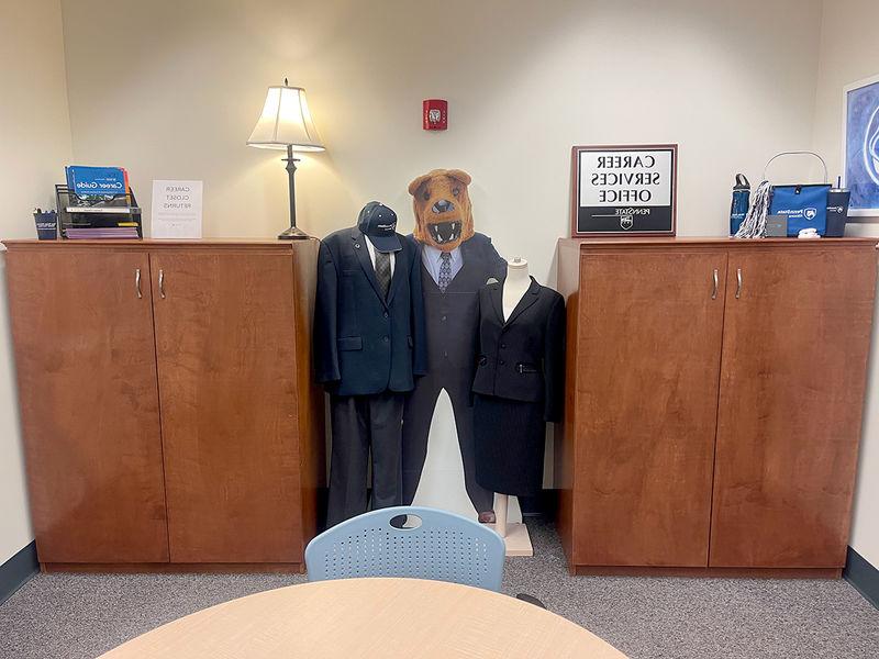 A man's and woman's suit on two dummies with a cardboard cutout of the Nittany Lion in a suit in the background between two large cupboards